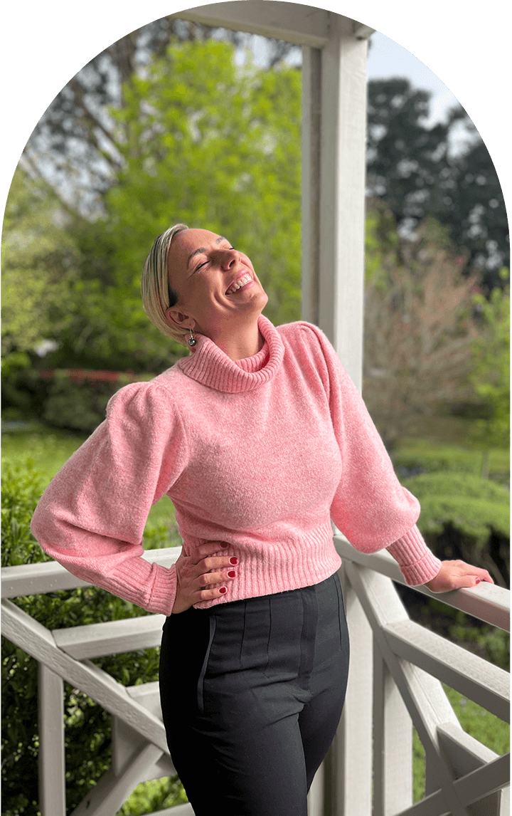 Rene Mariette, an SEO Copywriter based in Melbourne, standing in the sun in a pink jumper and smiling with her eyes closed.